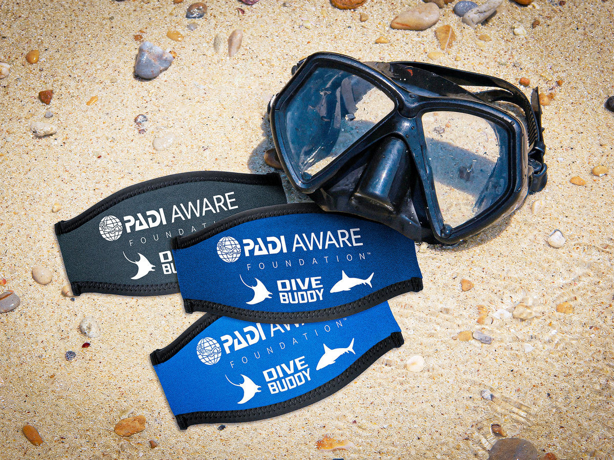 Three PADI AWARE Dive Buddy mask straps in black, navy and blue, beside a dive mask on sand