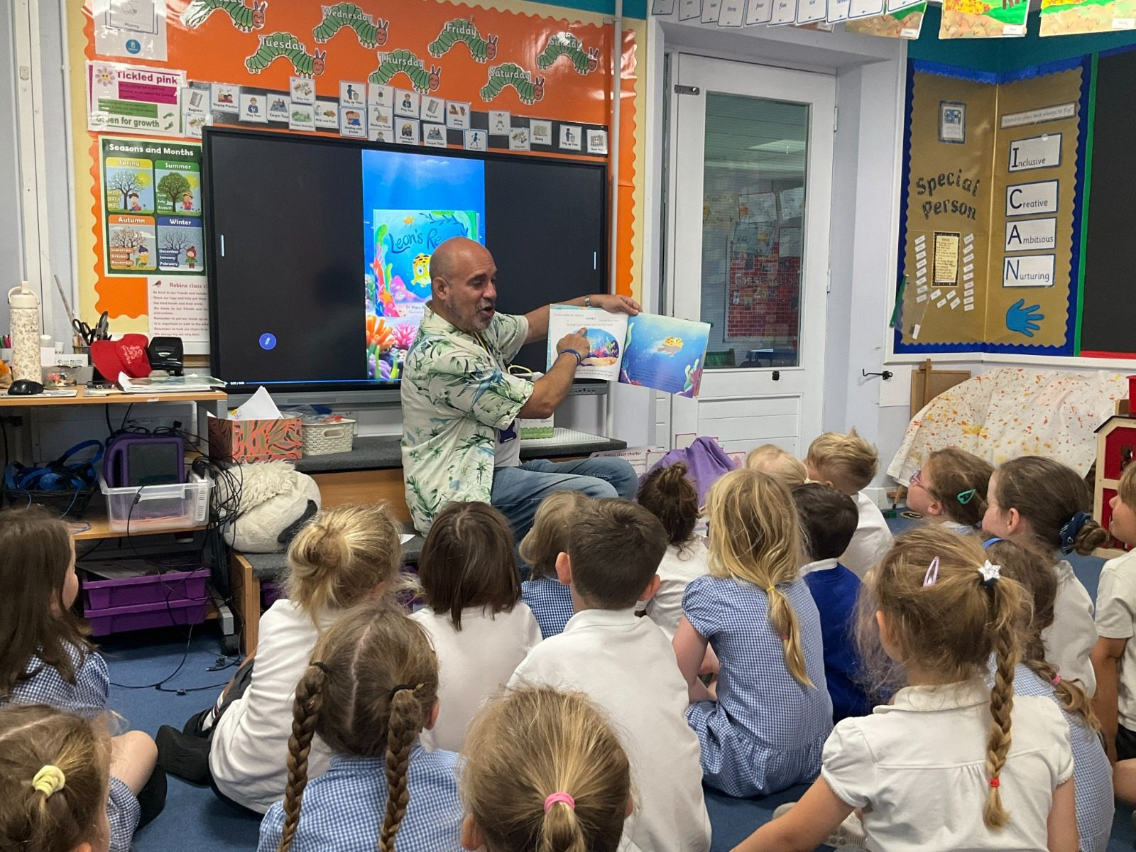 Dr Marc Abraham OBE reading Leon's Reef to a class of infant school pupils sitting on the floor